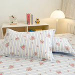 Cette image montre un lit avec des draps et taies d'oreiller blancs avec des fleurs roses sur un fond de rayures verticales. Il y a une lampe de table et une étagère sur le côté avec quelques livres et un jouet.