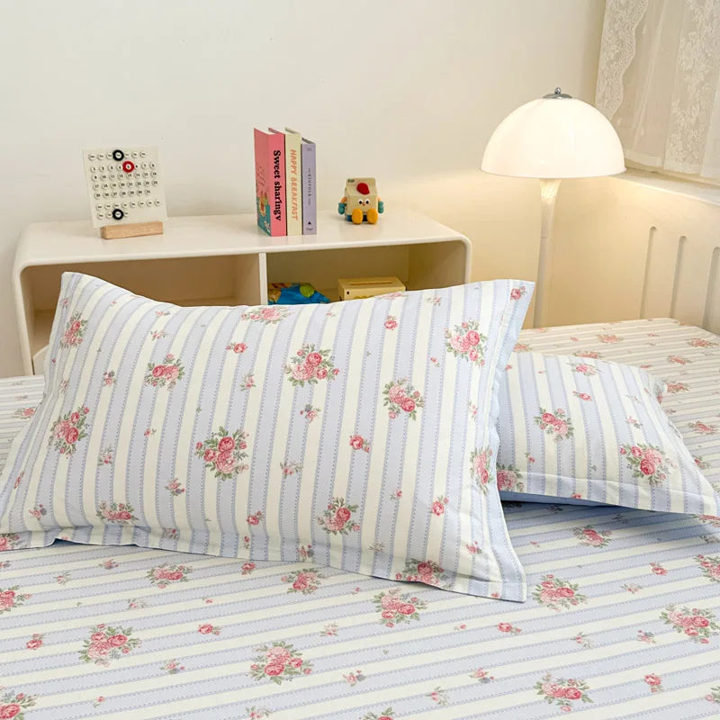 Cette image montre un lit avec des draps et taies d'oreiller blancs avec des fleurs roses sur un fond de rayures verticales. Il y a une lampe de table et une étagère sur le côté avec quelques livres et un jouet.