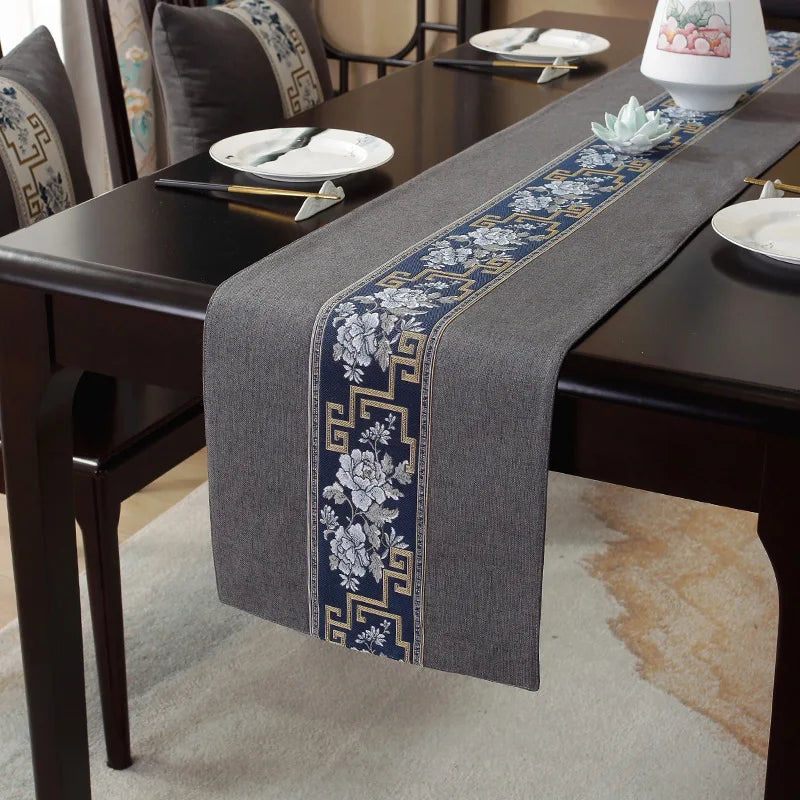 Chemin de table élégant avec des motifs floraux bleus et dorés sur fond gris, posé sur une table sombre dans un décor asiatique.