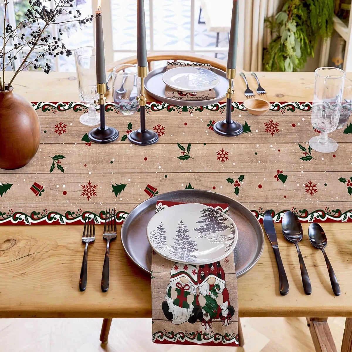 Une table de fête élégante avec des décorations de Noël, des bougies, des verres et des couverts en métal noir sur une nappe à motifs de flocons de neige et de branches de houx.