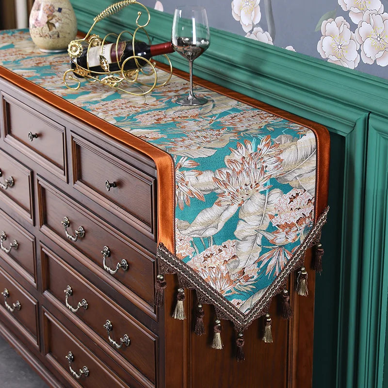 Un magnifique meuble de rangement vintage avec un chemin de table décoratif orné de motifs floraux et de glands, complété par un étagère verte, un verre de vin et des objets décoratifs, créant une atmosphère chaleureuse et élégante.