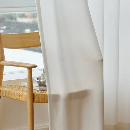 Voilage blanc soufflé par le vent, posé devant une grande fenêtre avec une chaise en bois au style scandinave