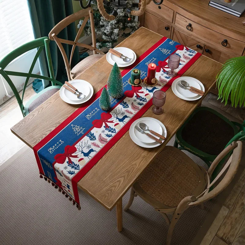 La table de salle à manger est joliment décorée pour les fêtes, avec un chemin de table rouge et bleu orné de motifs de feuilles et de rubans. Une petite figurine d'arbre de Noël complète la décoration festive.