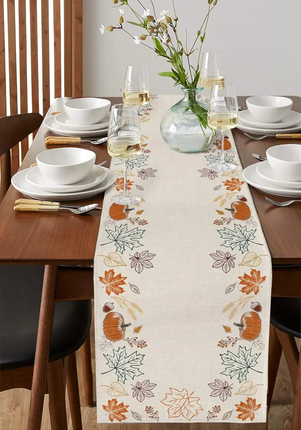Une table de salle à manger joliment dressée avec de la vaisselle blanche, des verres à vin, et une décoration automnale de feuilles et de citrouilles sur un chemin de table.