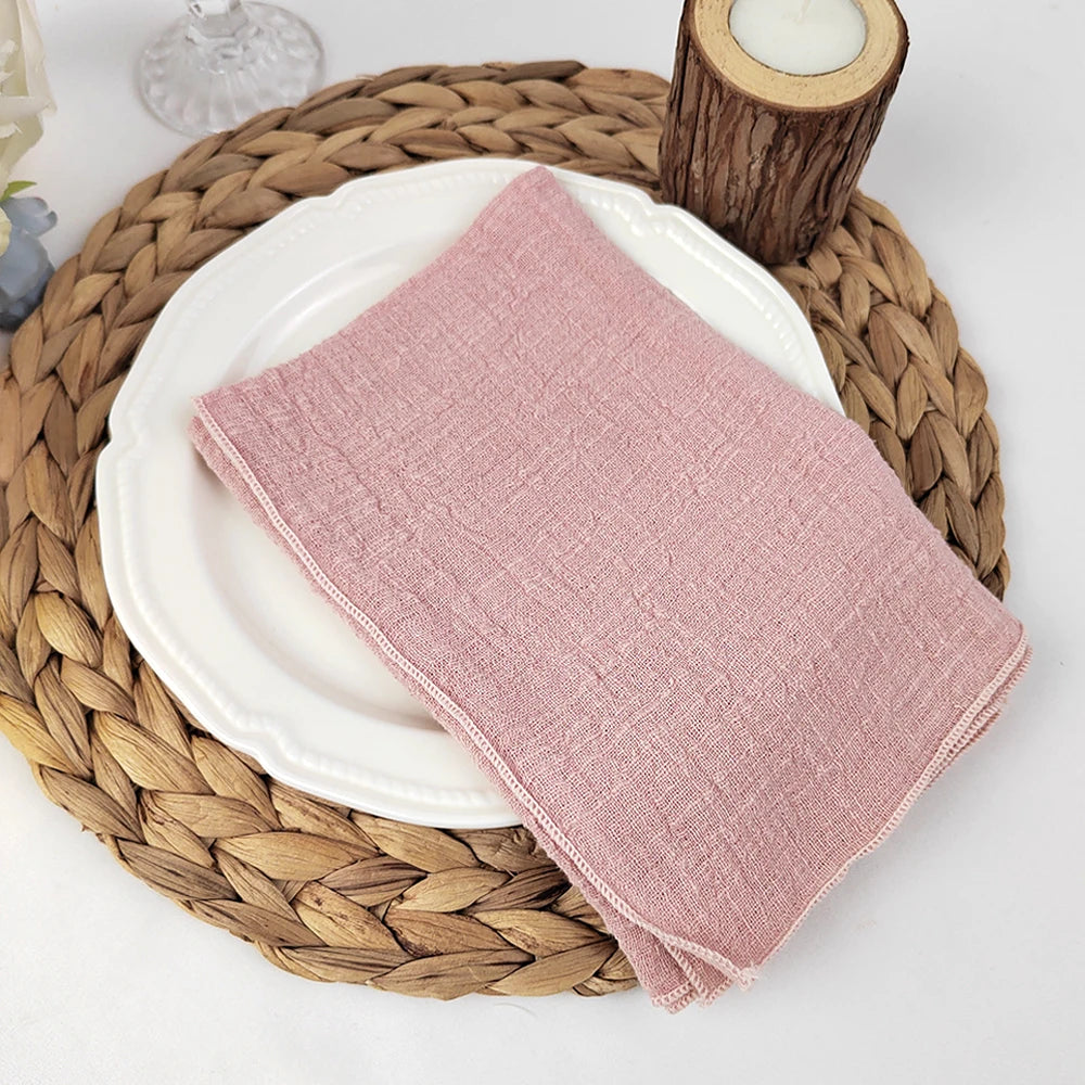 Serviette en lin rose sur une assiette blanche, posée sur un dessous de plat en fibres tressées, avec un support bougeoir en bois à côté.