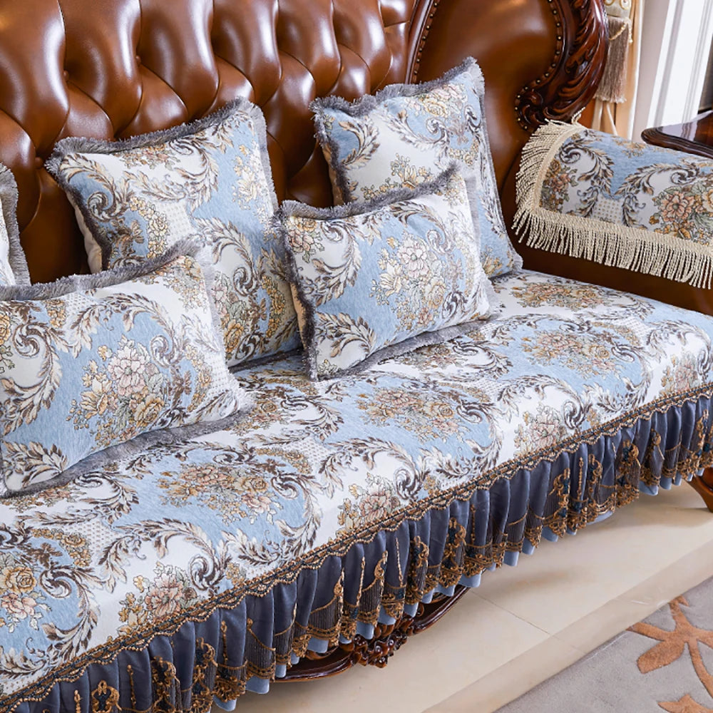 Un ensemble de literie de luxe avec des motifs floraux ornés de franges et de surpiqûres dans des tons gris et dorés, sur un canapé en cuir brun.