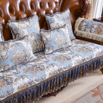 Un ensemble de literie de luxe avec des motifs floraux ornés de franges et de surpiqûres dans des tons gris et dorés, sur un canapé en cuir brun.