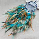 Un capteur de rêves en plumes bleu-vert et orange avec des motifs à pois, suspendu sur un fond gris clair.