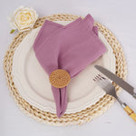 Une serviette de table rose pâle avec un nœud ornemental en tresses naturelles, posée sur un dessous de plat tressé, accompagnée d'une rose blanche.