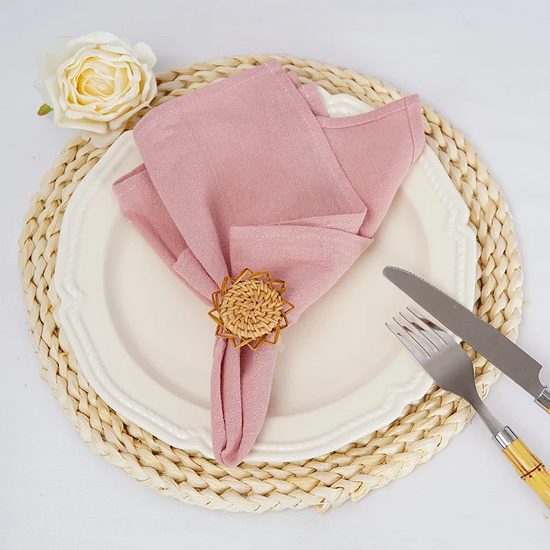 Un élégant couvert de table, composé d'une assiette blanche, d'une serviette rose pâle pliée avec un ornement doré, et d'une rose jaune sur un tapis tressé.