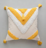 Coussin décoratif moelleux en tissu blanc avec des triangles géométriques jaunes et des pompons assortis.