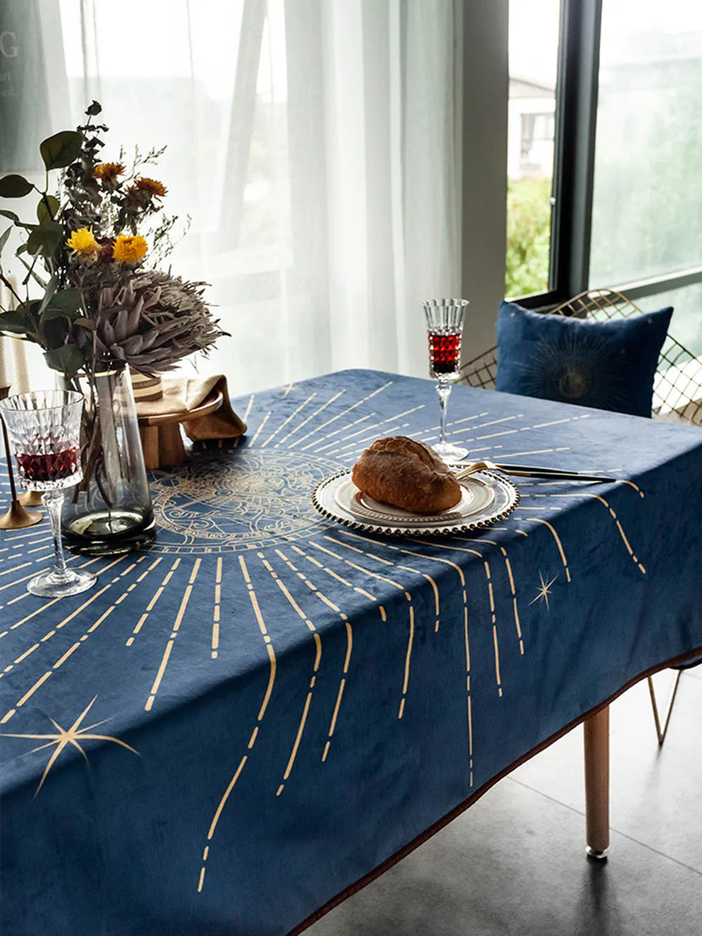 Une table élégante avec un centre-de-table floral, un verre de cristal et un petit pain sur une assiette, le tout rehaussé par une nappe bleu marine ornée de motifs dorés.