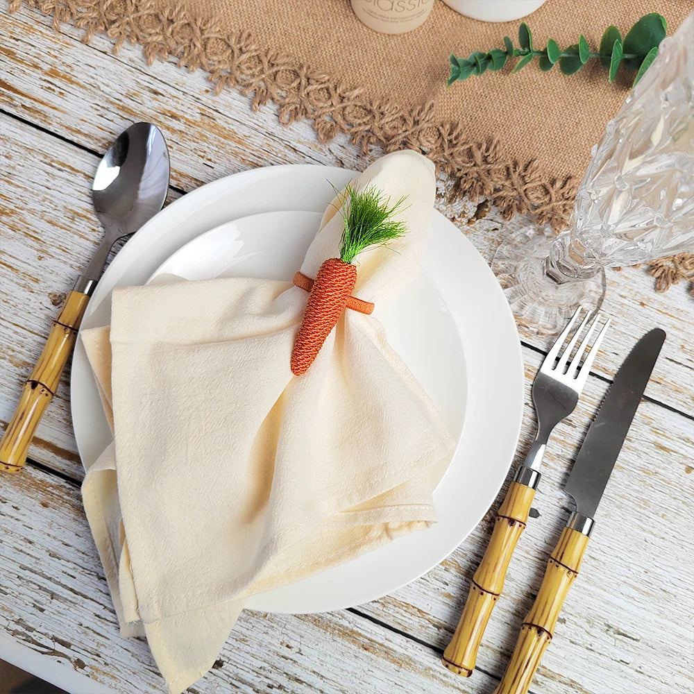Une assiette blanche décorée d'une serviette beige enroulée et ornée d'un petit carotte verte, sur un fond de bois clair avec des couverts en métal doré.