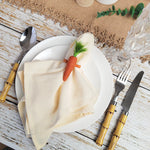 Une assiette blanche décorée d'une serviette beige enroulée et ornée d'un petit carotte verte, sur un fond de bois clair avec des couverts en métal doré.