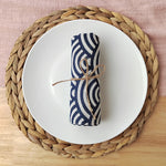 Serviette de table blanche et bleue rayée, attachée avec une ficelle, posée sur un napperon rond en fibres tressées.