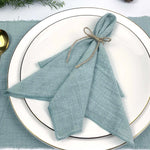 Des serviettes de table bleu-vert nouées avec de la ficelle naturelle, disposées sur une assiette blanche aux bordures dorées, entourées de branches de feuillage vert.