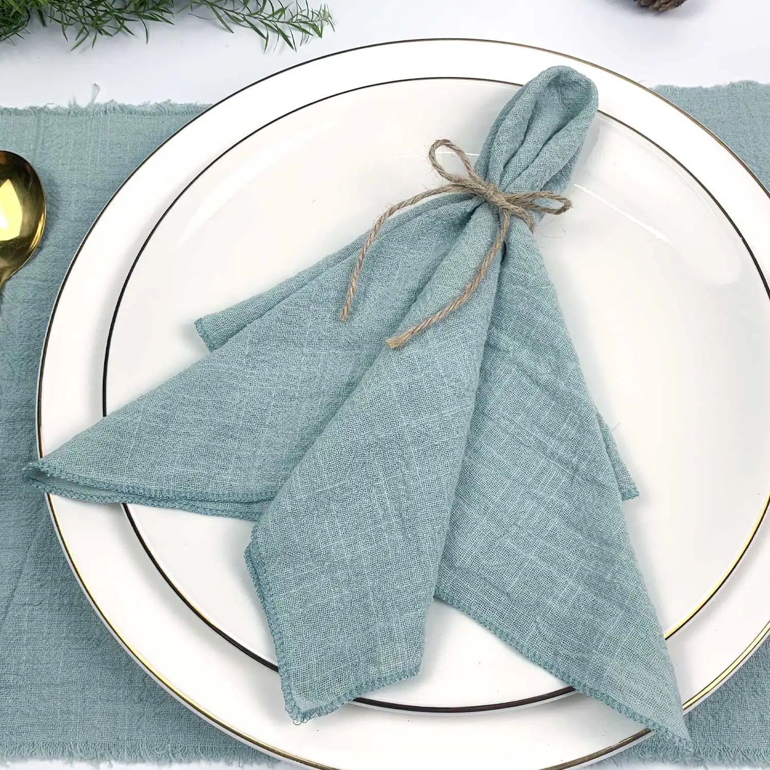 Des serviettes de table bleu-vert nouées avec de la ficelle naturelle, disposées sur une assiette blanche aux bordures dorées, entourées de branches de feuillage vert.