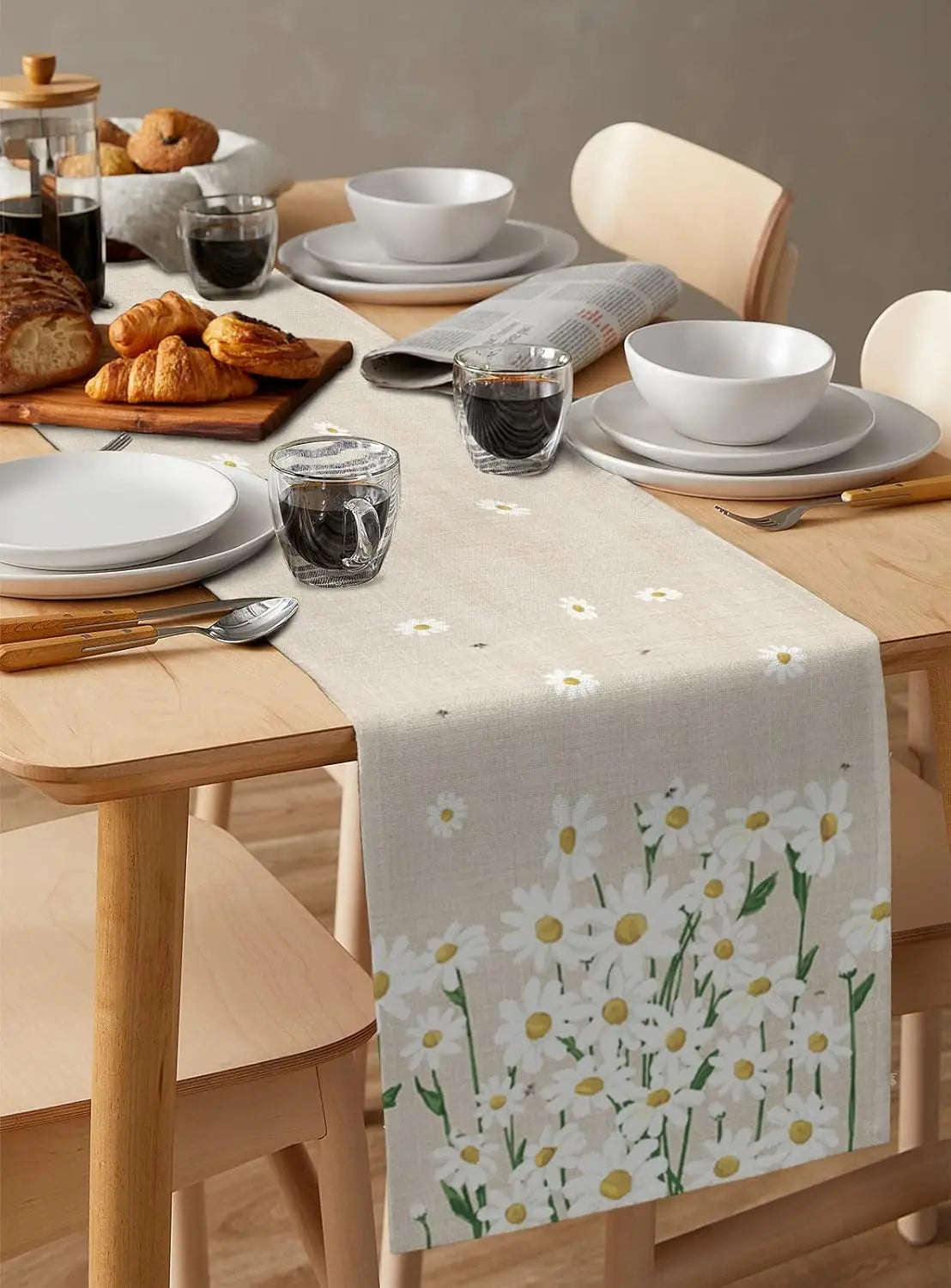 Une table de petit-déjeuner avec des pains frais, des bols blancs, des verres et une nappe ornée de motifs de marguerites blanches.