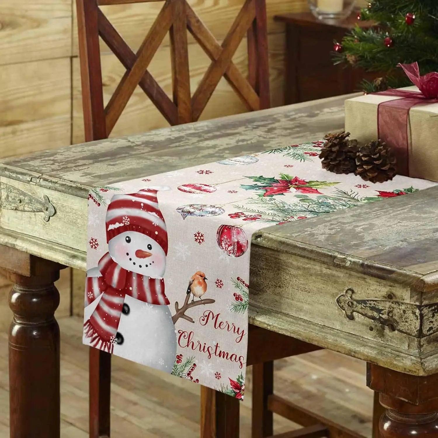 Une composition festive avec un bonhomme de neige sur un chemin de table décoré de pommes de pin et d'éléments de Noël, sur un meuble ancien en bois.