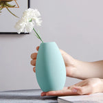 Un élégant vase vert menthe de forme ovale posé sur une surface grise, avec une fleur blanche immaculée à l'intérieur, créant une composition minimaliste et raffinée.