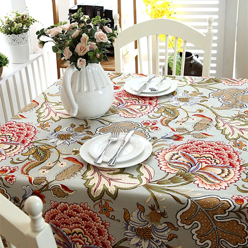Cette image présente un beau décor de table avec une nappe florale colorée, une théière blanche et des assiettes blanches posées sur la table. Des fleurs fraîches dans un vase blanc ajoutent une touche de nature à cette scène d'intérieur chaleureuse et accueillante.