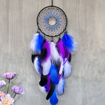 Un attrape-rêves orné de plumes colorées, notamment bleu et violet, suspendu sur un mur gris, avec des fleurs en arrière-plan.