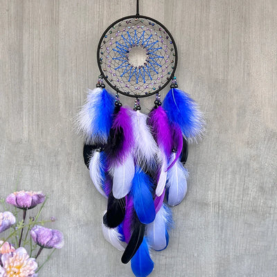Un attrape-rêves orné de plumes colorées, notamment bleu et violet, suspendu sur un mur gris, avec des fleurs en arrière-plan.