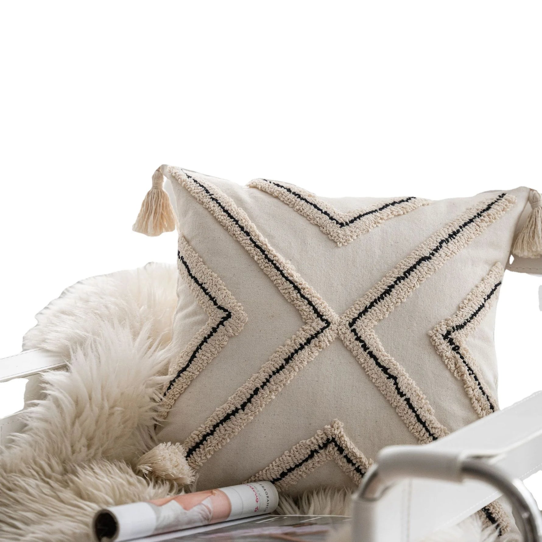Coussin décoratif en coton beige orné de motifs graphiques et de pompons, posé sur une couverture en fourrure blanche, aux côtés de magazines.