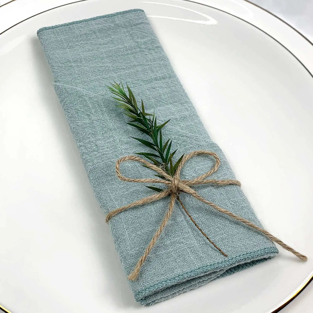 Une serviette de cuisine gris-vert soigneusement pliée, ornée d'un brin de romarin lié par une ficelle naturelle, sur une assiette blanche.