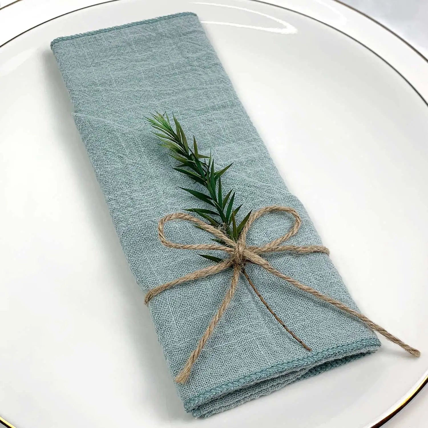 Une serviette de cuisine gris-vert soigneusement pliée, ornée d'un brin de romarin lié par une ficelle naturelle, sur une assiette blanche.