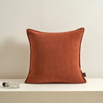 Coussin décoratif en tissu d'un ton orangé terre cuite posé sur une surface blanche.