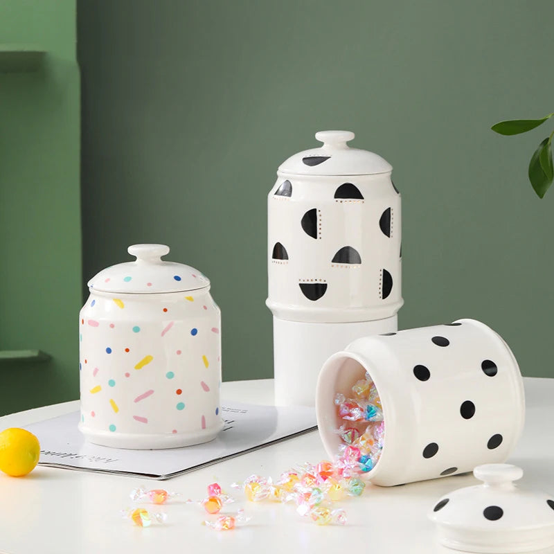 Des boîtes de rangement en céramique colorées et ponctuées de motifs géométriques, contenant des bonbons et d'autres éléments décoratifs, sur un fond vert pastel.