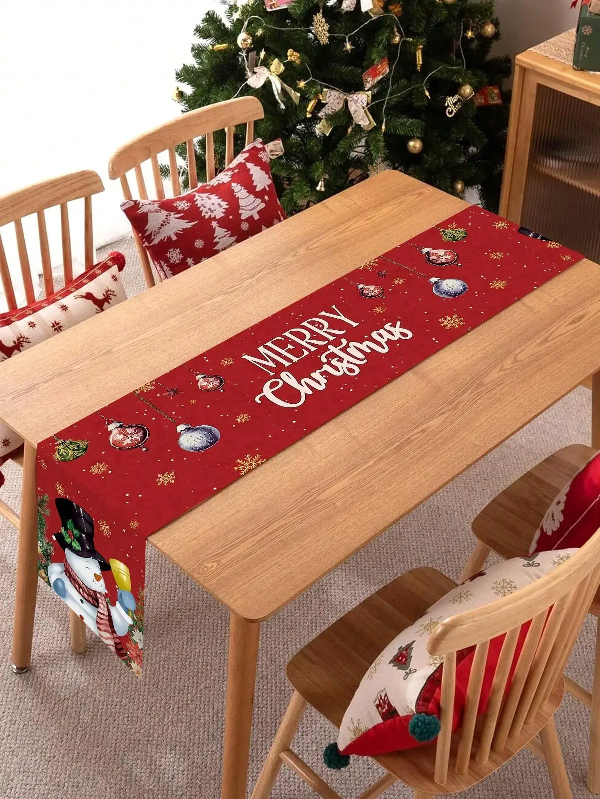 Table de salle à manger en bois avec un chemin de table rouge décoré de motifs de Noël et de la mention "Joyeux Noël", chaises en bois, et un sapin de Noël décoré à l'arrière-plan.