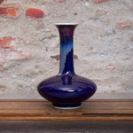Vase en céramique de style moderne, de couleur bleu foncé avec des touches de bleu plus pâle, posé sur une surface en bois, devant un mur de pierre.