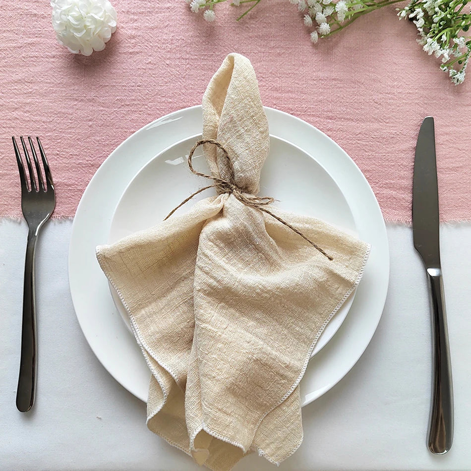Une assiette blanche avec des couverts en acier et un linge de table beige noué artistiquement, sur un fond rose pastel.