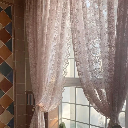 Voilage en dentelle rose clair à motifs floraux délicats, attaché partiellement pour laisser entrer la lumière naturelle dans une pièce carrelée chaleureuse.
