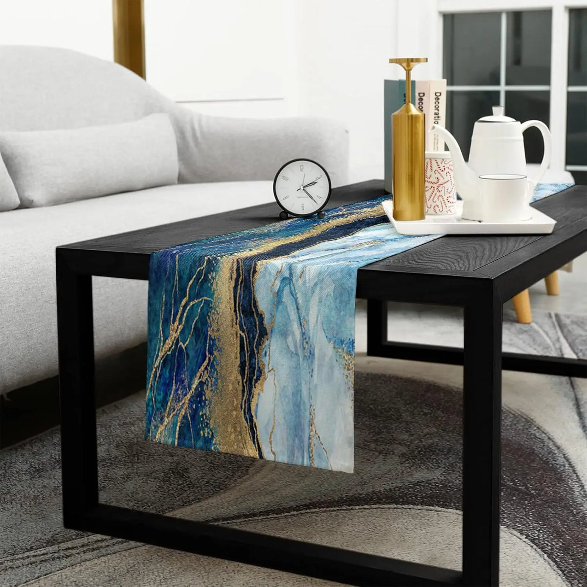 Table basse en bois noir avec un chemin de table en motif marbré bleu et doré, posée sur un tapis gris dans un salon lumineux avec canapé blanc.