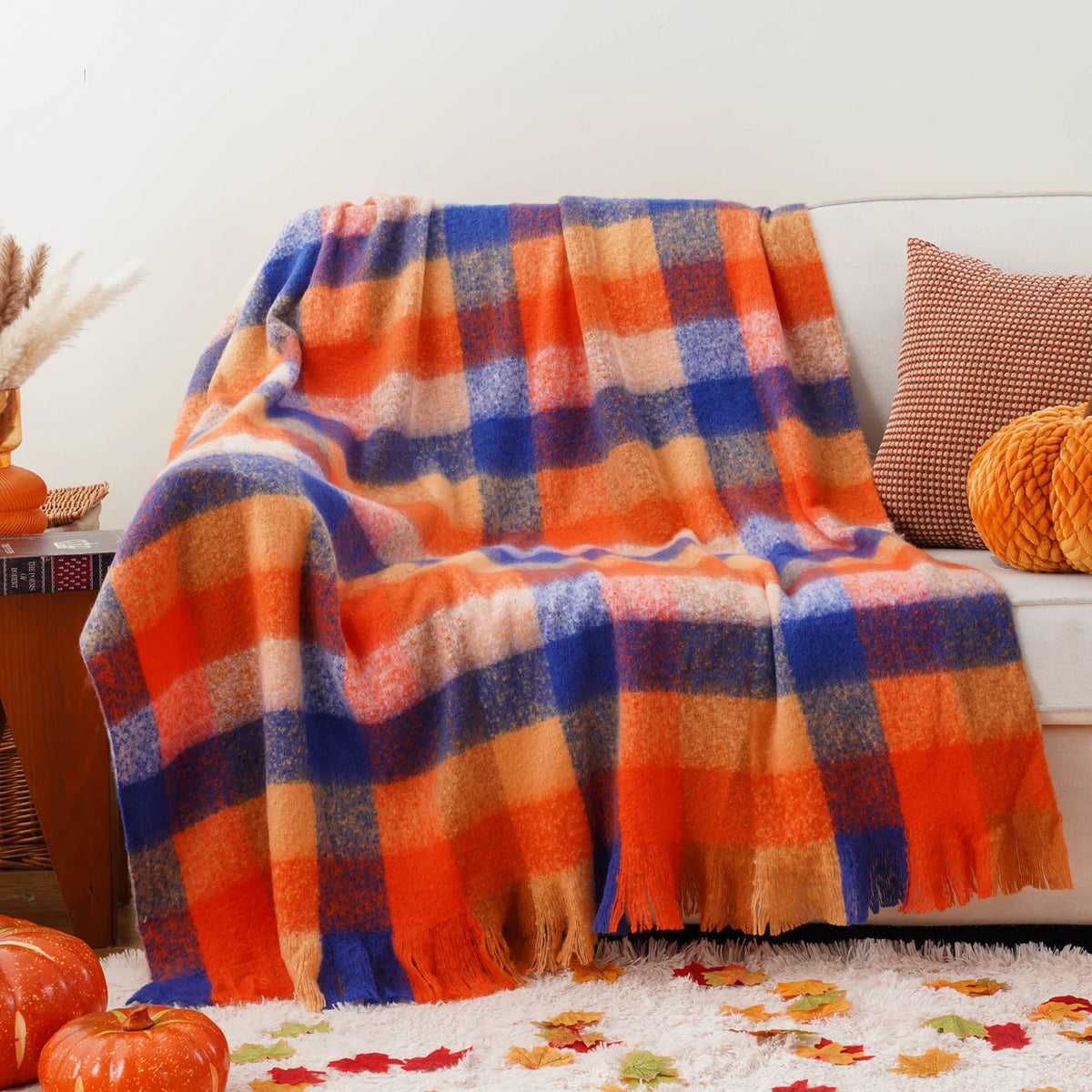 Cette image montre une couverture en laine multicolore aux motifs carreaux bleus et oranges, posée sur un canapé avec des coussins colorés. Elle crée une ambiance chaleureuse et automnale, avec des feuilles mortes au sol, rappelant une atmosphère cocooning.