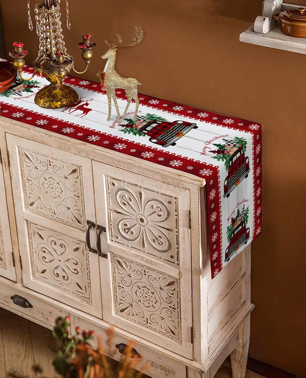 Meuble de rangement décoré de motifs floraux sculptés, recouvert d'un linge de table rouge festif orné de flocons de neige et d'un véhicule vintage, accompagné de figurines métalliques de cerfs dorés.
