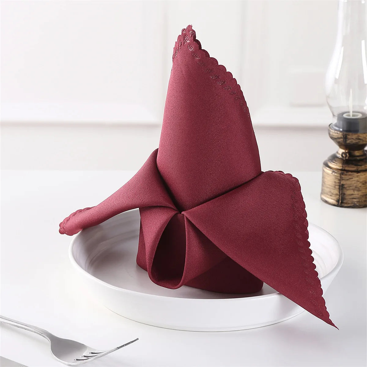 Serviette de table en tissu rouge bordeaux avec un bord festonné et un noeud décoratif, posée sur une assiette blanche avec des couverts à côté.