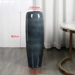 Un grand vase en céramique de couleur gris foncé, mesurant 65 cm de hauteur et 15 cm de diamètre, posé sur un parquet brun clair dans un environnement lumineux.