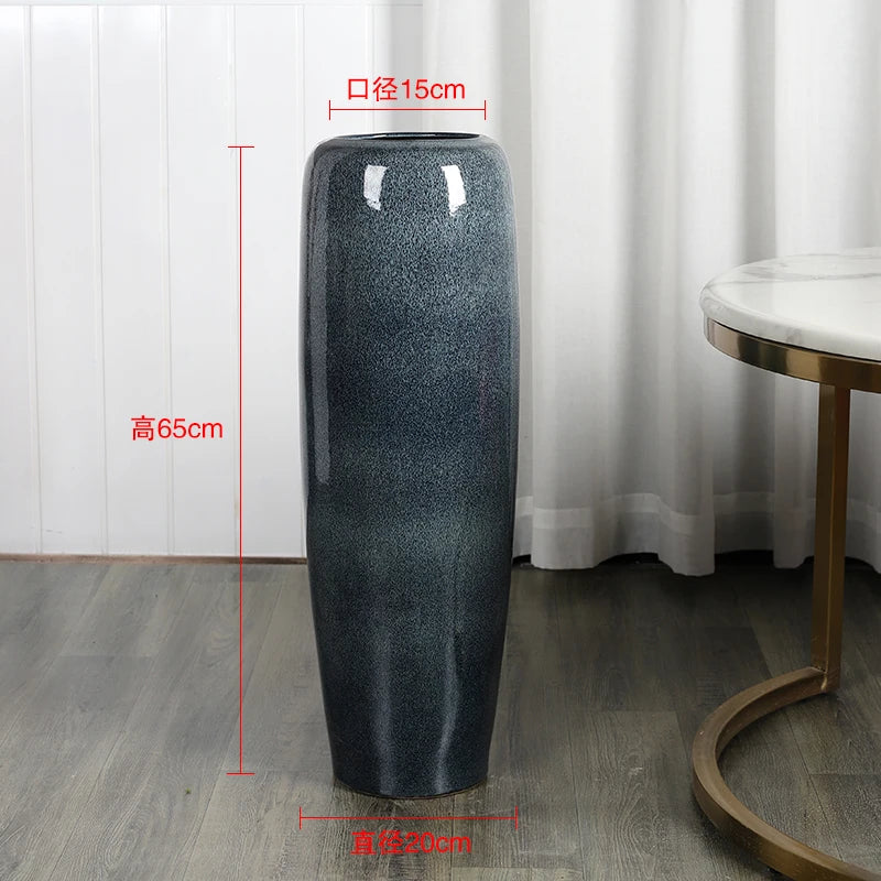 Un grand vase en céramique de couleur gris foncé, mesurant 65 cm de hauteur et 15 cm de diamètre, posé sur un parquet brun clair dans un environnement lumineux.