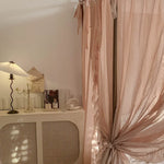 Un intérieur élégant et douillet avec des rideaux rose pâle, des objets décoratifs sur un meuble et une lampe de table vintage, créant une atmosphère chaleureuse et apaisante.