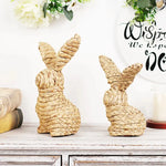 Deux adorables lapins décorés en osier, posés sur une table vintage avec des fleurs blanches, créant une scène printanière chaleureuse et bucolique.
