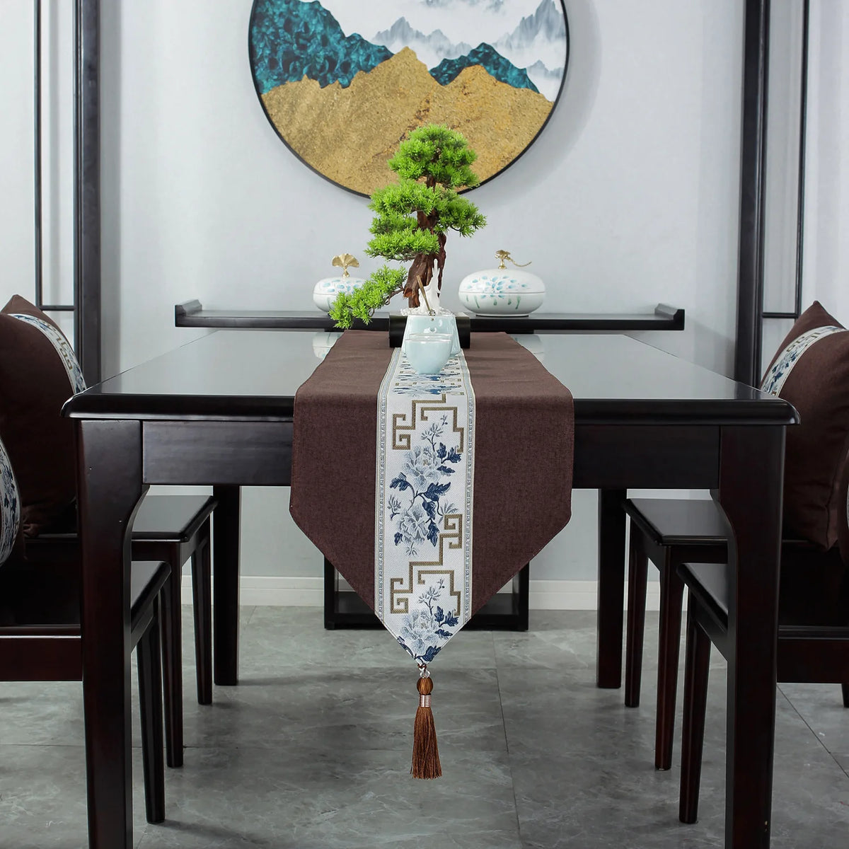 Cette image montre une table à manger avec un magnifique décor japonisant, comprenant un chemin de table en tissu brun orné de motifs bleus et dorés, ainsi qu'un bonsaï placé au centre de la table, le tout encadré par une œuvre d'art murale aux couleurs chaudes.