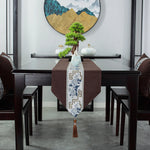 Cette image montre une table à manger avec un magnifique décor japonisant, comprenant un chemin de table en tissu brun orné de motifs bleus et dorés, ainsi qu'un bonsaï placé au centre de la table, le tout encadré par une œuvre d'art murale aux couleurs chaudes.