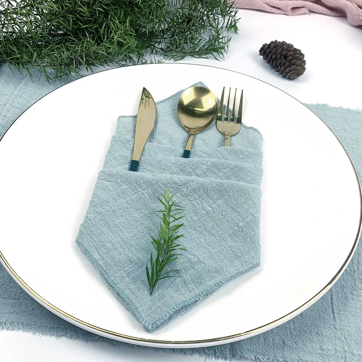 Une élégante configuration de table, avec une serviette de table en lin bleu tendre, une fourchette et une cuillère dorées, et une touche naturelle apportée par un rameau de feuillage vert.