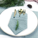 Une élégante configuration de table, avec une serviette de table en lin bleu tendre, une fourchette et une cuillère dorées, et une touche naturelle apportée par un rameau de feuillage vert.