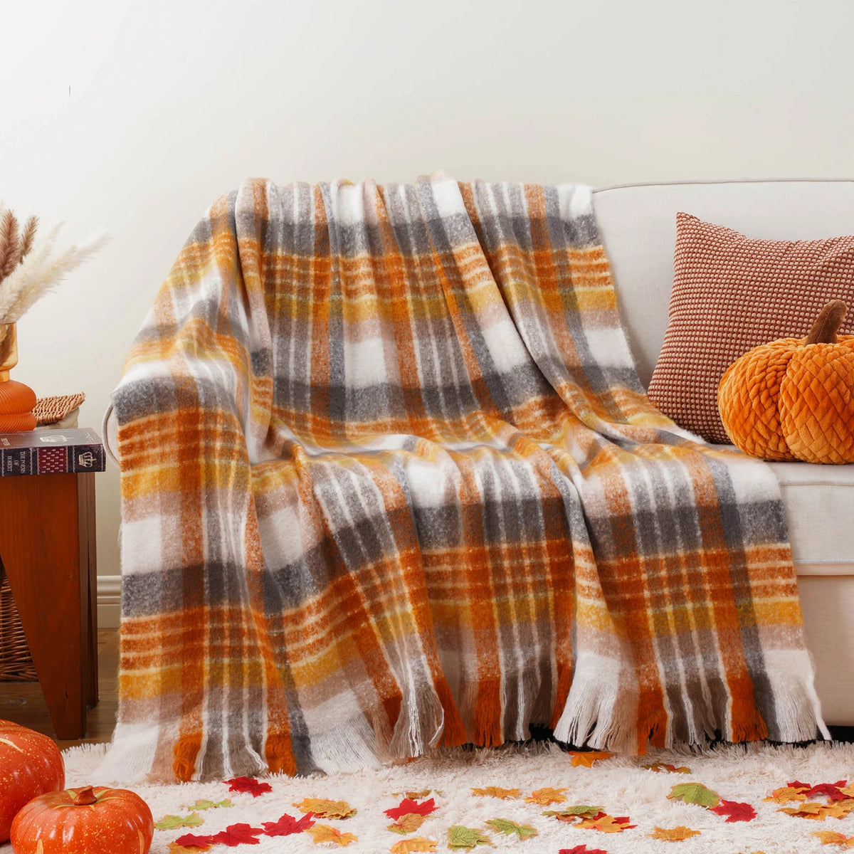 Une couverture à carreaux en gris et orange, aux accents automnaux, reposant sur un canapé avec des coussins colorés et des feuilles d'automne éparpillées.