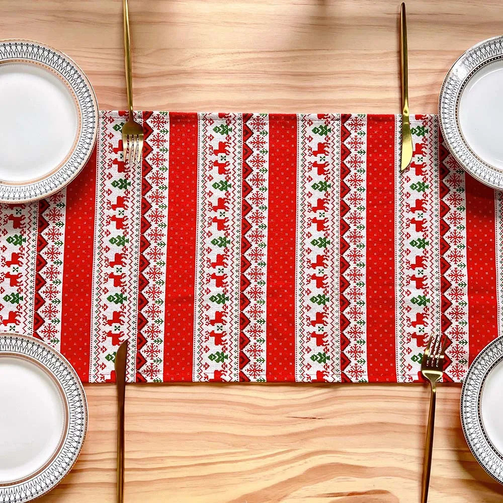 Nappe de table festive, ornée de motifs traditionnels rouges et verts, sur un fond en bois clair, entourée d'assiettes blanches élégantes.